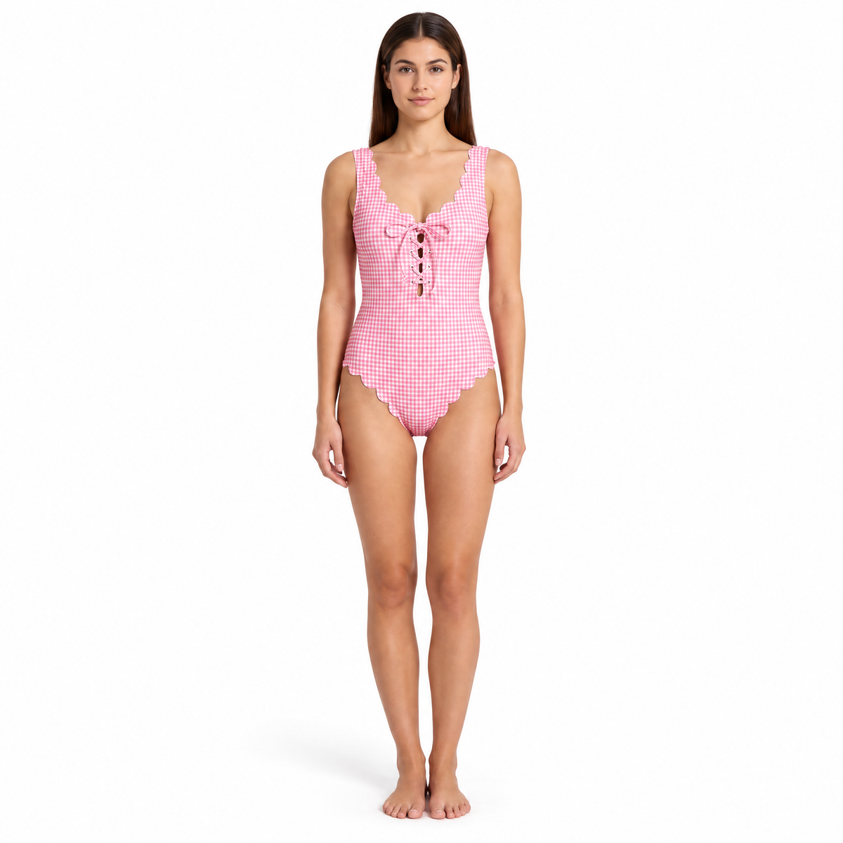 A woman modeling a pink and white gingham one-piece swimsuit with scalloped edges and a lace-up front, standing against a plain white background.