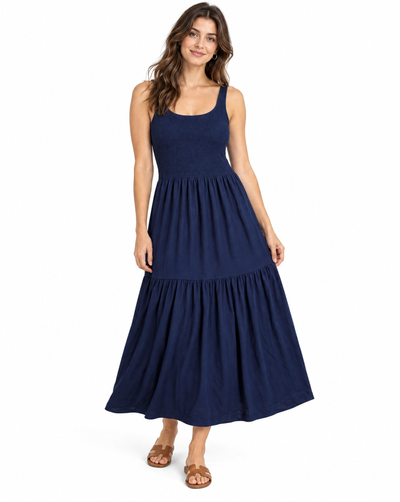 A woman modeling a sleeveless navy blue maxi dress with a tiered skirt, paired with brown leather sandals, against a plain white background.